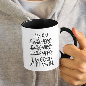 Può includere: Tazza in ceramica bianca con interno e manico neri. La tazza ha il testo nero "I'M AN ENGINEER I'M GOOD WITH MATH". La parola "engineer" è stata cancellata più volte.