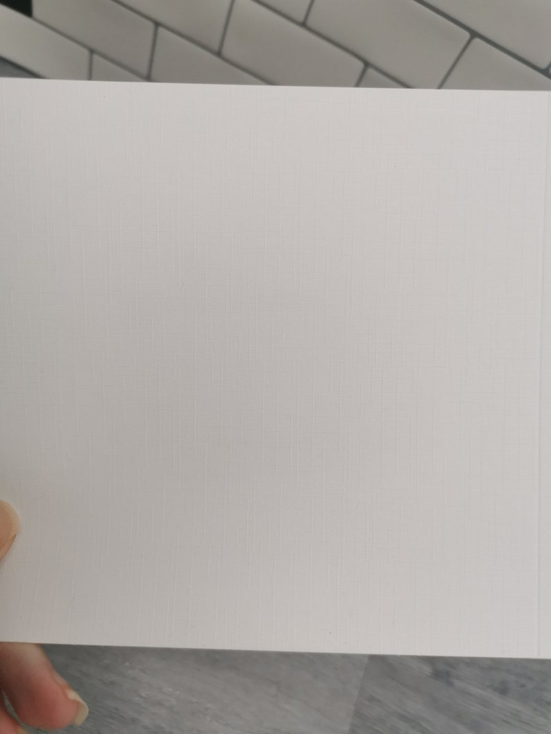 May include: A close-up of a white, textured paper with a subtle grid pattern. The paper is held by a hand, and the background features a white and gray tiled surface.