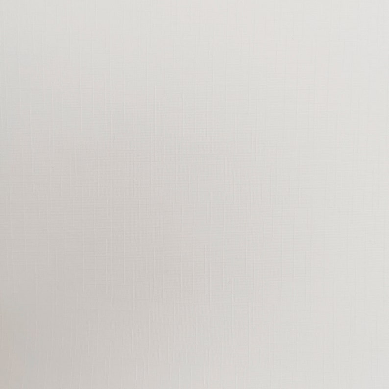 May include: A close-up of a white, textured fabric. The fabric has a subtle grid pattern, with vertical and horizontal lines creating a woven appearance. The color is a soft, off-white, providing a neutral background.