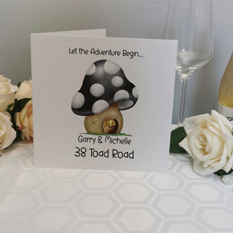 May include: A folded white card with a whimsical mushroom house illustration. The mushroom has a black and white spotted cap and a brown base with a door and window. Text reads "Let the Adventure Begin..." and names and address.