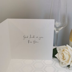May include: A white greeting card with the message "Good Luck in your New Home" in elegant script. A champagne flute and a white rose are in the background, suggesting a celebratory theme.