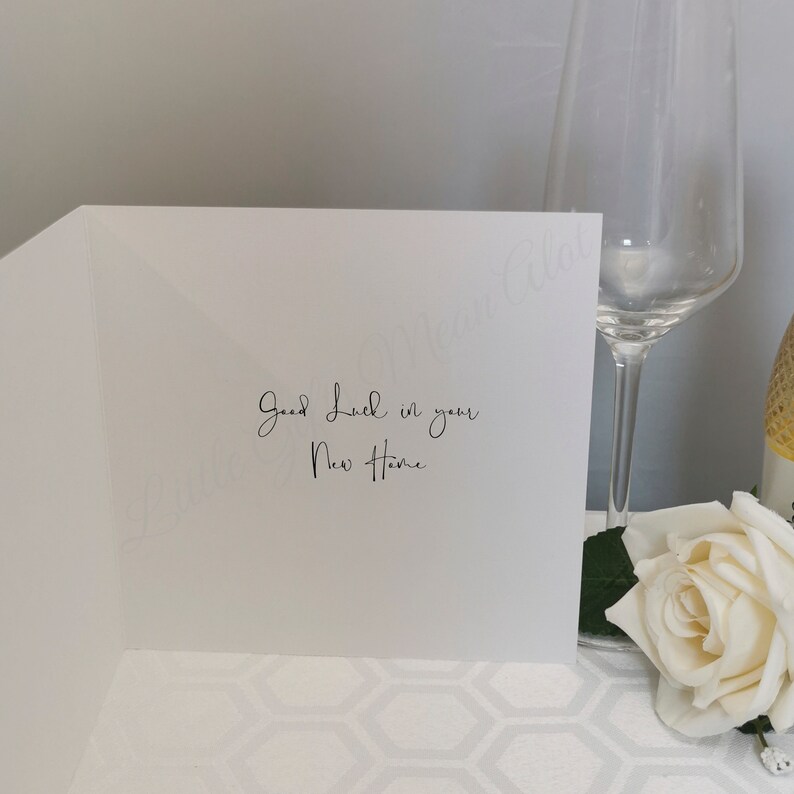 May include: A white greeting card with the message "Good Luck in your New Home" in elegant script. A champagne flute and a white rose are in the background, suggesting a celebratory theme.