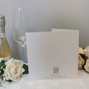 May include: A folded white greeting card with a QR code and text, next to a champagne bottle and a tall wine glass. A white rose and other flowers are in the foreground. The background is a light gray wall.