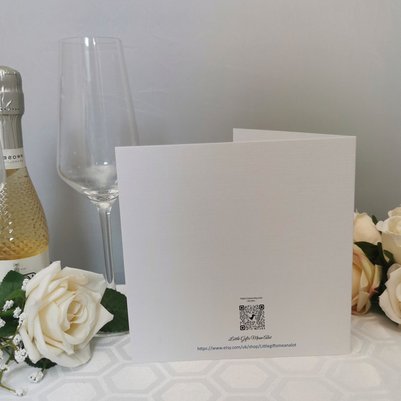 May include: A folded white greeting card with a QR code and text, next to a champagne bottle and a tall wine glass. A white rose and other flowers are in the foreground. The background is a light gray wall.