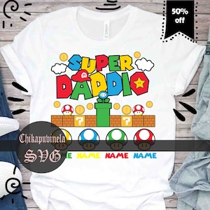 Può includere: T-shirt bianco con un design grafico colorato con la scritta "SUPER DADDIO" in un font in stile videogioco. Il design include elementi come monete, funghi e una stella. La maglietta ha anche il testo "50% off".