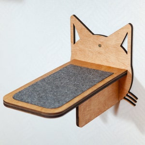 Pode incluir: Uma prateleira de gato de madeira em forma de cabeça de gato com uma almofada de feltro cinza no topo. A prateleira é montada em uma parede branca.