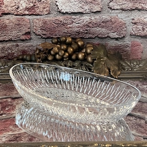 Puede incluir: Fuente ovalada de vidrio transparente con un diseño texturizado y acanalado. El plato se refleja en la superficie inferior. Un racimo de uvas y hojas decorativas de bronce se encuentran en el fondo contra una pared de ladrillos.