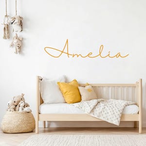 May include: A wall decal featuring the name Amelia in a cursive, golden-yellow script font. The decal is applied to a plain white wall above a light-coloured wooden daybed, which is decorated with white and yellow pillows and a patterned blanket.