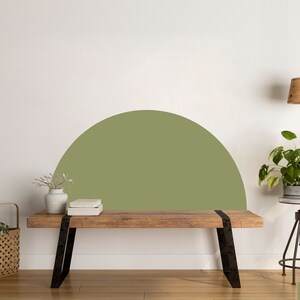 May include: A wooden bench with black metal legs sits in front of a white wall with a half-circle painted in olive green. A potted plant sits on a metal stool next to the bench. The word "Olive" is written in green at the bottom of the image.