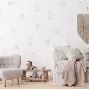 K&ouml;nnte beinhalten: Eine Wand mit einem sich wiederholenden Muster aus beigen Linien-Hasen-Aufklebern. Die Hasen sind in verschiedenen Posen dargestellt, darunter sitzend, liegend und stehend. Die Wand ist Teil eines Raumes mit einem hellen Sessel, einer Holzbank mit Pl&uuml;schtieren und einem Tagesbett mit Kissen und einem Makramee-Wandbehang.