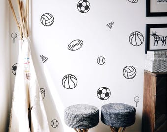 May include: A white wall decorated with black sports-themed decals, including basketballs, soccer balls, footballs, baseballs, volleyballs, golf balls, and badminton shuttlecocks. Two gray woven stools with wooden legs are in the foreground. A teepee is on the left.