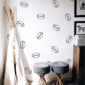 May include: A set of black American football wall decals featuring a simple line art design of a rugby-style ball. These individual stickers are arranged in a scattered pattern across a white wall, providing a decorative accent for a room.