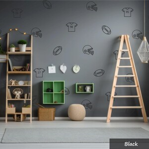 May include: A dark grey wall featuring black line art decals of football helmets, jerseys with numbers, and footballs. The wall includes a wooden shelving unit, a green four-compartment wall shelf, a small green shelf, and a wooden ladder.