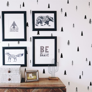 May include: A wall display featuring three black-framed prints with woodland-themed graphics and text, including a bear, a fox, and a mountain. The wall is decorated with black pine tree decals. A wire cactus sculpture and two patterned boxes sit below.