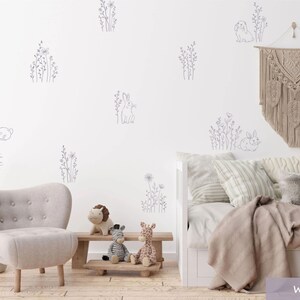 K&ouml;nnte beinhalten: Ein Set von Wandaufklebern mit grauen Strichzeichnungen von Kaninchen und Wildblumen auf einer wei&szlig;en Wand. Die Szene umfasst einen hellgrauen Sessel, eine Holzbank mit Pl&uuml;schtieren und ein Tagesbett mit neutralen Kissen.