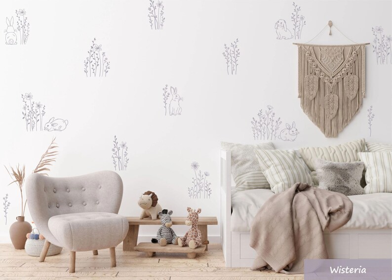 K&ouml;nnte beinhalten: Ein Set von Wandaufklebern mit grauen Strichzeichnungen von Kaninchen und Wildblumen auf einer wei&szlig;en Wand. Die Szene umfasst einen hellgrauen Sessel, eine Holzbank mit Pl&uuml;schtieren und ein Tagesbett mit neutralen Kissen.