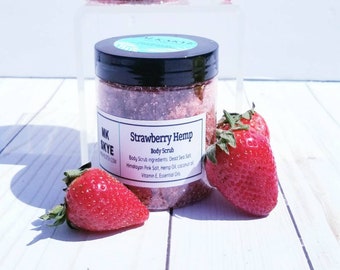 Può includere: Due barattoli di vetro di scrub corpo alla fragola e canapa con coperchi neri. I barattoli sono su una superficie di legno bianco con fragole fresche.