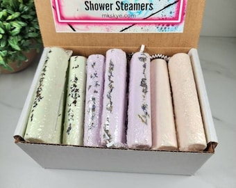 May include: A white box with a brown lid containing a set of eight shower steamers. The lid has a label that reads "MK Skye Bath & Body Shower Steamers mksky.com". The steamers are in three colors: green, purple, and pink.