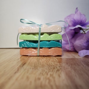 May include: A stack of four handmade soap bars wrapped in cellophane and tied with a light blue ribbon. The soap bars are in shades of green, peach, and white.