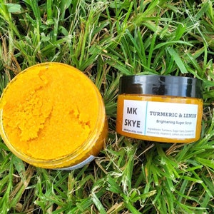 May include: A glass jar filled with a bright orange turmeric and lemon sugar scrub. The jar is labeled "MK SKYE" and "TURMERIC & LEMON Brightening Sugar Scrub". The ingredients are listed as "Turmeric, Sugar Cane, Coconut Oil, Almond Oil, Vitamin E, Lemon zest, essential oil".