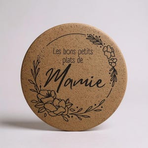 Può includere: Un sottobicchiere rotondo in sughero con la scritta "Les bons petits plats de Mamie" in nero. Il sottobicchiere è decorato con un motivo a ghirlanda floreale. Il sottobicchiere è di colore marrone chiaro.