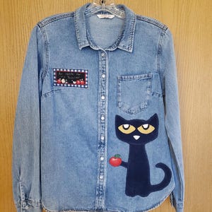 May include: Light blue denim button-up shirt with long sleeves. Features a black cat graphic holding a red apple on the front. A patch with the text "An apple for my teacher" is on the left chest. The shirt is hanging on a hanger.