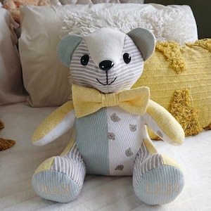 May include: A patchwork teddy bear made from various fabrics in shades of light blue, yellow, and white with a striped and bear-patterned design. It features a yellow bow tie, black eyes, and the name DEAN and date 1.2.24 embroidered on its feet.