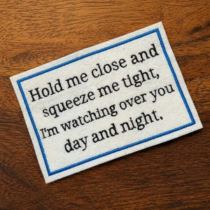 May include: A rectangular white fabric patch with a blue embroidered border. The patch features black embroidered text that reads: Hold me close and squeeze me tight, I'm watching over you day and night. The design is clean and simple.