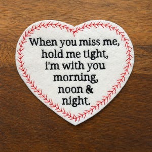 May include: A white heart-shaped felt patch with red baseball-style stitching along the edge. The black embroidered text reads: When you miss me, hold me tight, I'm with you morning, noon & night. The patch has a clean, flat, and symmetrical design.