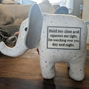 May include: A white fabric elephant toy featuring a pattern of small multicoloured stars. A rectangular patch on the side displays the black text: Hold me close and squeeze me tight, I'm watching over you day and night. The elephant has grey ears.