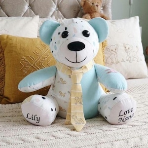 May include: A plush teddy bear made from light blue and white fabric with a patterned design featuring small animal illustrations. The bear wears a yellow striped necktie and has the names Lily and Love Nana embroidered on its feet.