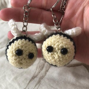 May include: Two handmade crochet bee keyrings. Each bee has a cream-coloured body with a black stripe, black eyes, and white antennae. The keyrings have silver-coloured metal rings and chains.