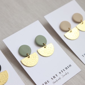 Puede incluir: Primer plano de pendientes hechos a mano sobre tarjetas blancas. Cada par presenta un perno circular en varios colores, conectado a un semicírculo texturizado de color dorado. Las tarjetas muestran "THE ART STUDIO Handmade Jewelry".