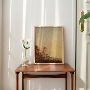 Puede incluir: Una impresi&oacute;n enmarcada de una pintura que representa un paisaje con palmeras en una luz c&aacute;lida y dorada. La impresi&oacute;n est&aacute; colgada en una pared sobre una mesa auxiliar de madera con un jarr&oacute;n de flores blancas.
