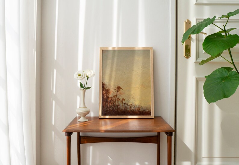 Puede incluir: Una impresi&oacute;n enmarcada de una pintura que representa un paisaje con palmeras en una luz c&aacute;lida y dorada. La impresi&oacute;n est&aacute; colgada en una pared sobre una mesa auxiliar de madera con un jarr&oacute;n de flores blancas.