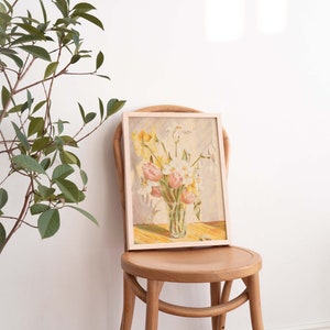 May include: A framed painting of a bouquet of yellow, white, and pink flowers in a glass vase. The painting is on a wooden chair with a light wood finish.