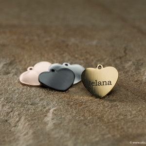 Könnte beinhalten: Vier herzförmige Metalletiketten, zwei silber, eines schwarz und eines gold. Das goldene Etikett trägt den Namen "Belana" eingraviert.