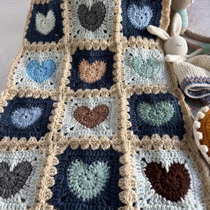 May include: A handmade crochet blanket featuring a grid of square motifs, each containing a central heart shape. The hearts and square borders are worked in various shades of blue, beige, brown, and sage green yarn, joined by beige yarn.