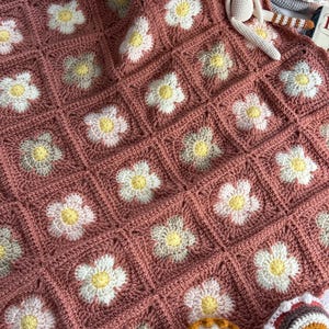 May include: A crochet blanket composed of square motifs featuring white and light grey flower petals with yellow centres, all joined by a dusty rose yarn. A small crocheted doll and two crescent-shaped toys are placed on the blanket surface.