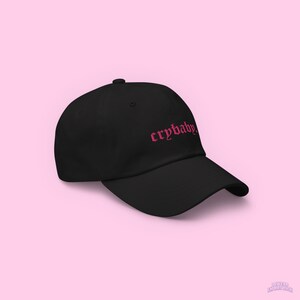 Puede incluir: Una gorra de b&eacute;isbol negra con un "crybaby" bordado en rosa en una fuente g&oacute;tica.