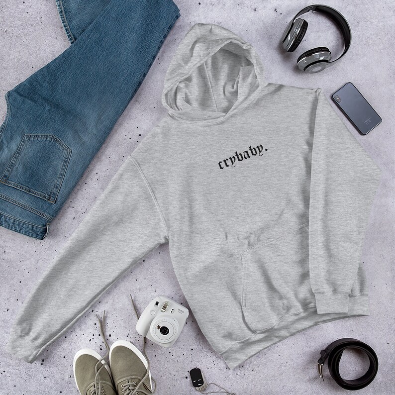 May include: Grey hooded sweatshirt with the text "crybaby." in black gothic font. The sweatshirt is laid flat on a grey textured surface with a pair of jeans, a camera, headphones, and a belt.