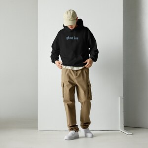 May include: A black hooded sweatshirt with the text "ghost boy" embroidered in blue on the front. The model is wearing the sweatshirt with beige cargo pants and a beige baseball cap.