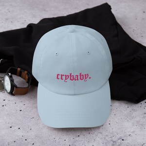 Puede incluir: Una gorra de b&eacute;isbol azul claro con el texto "crybaby" bordado en rosa.