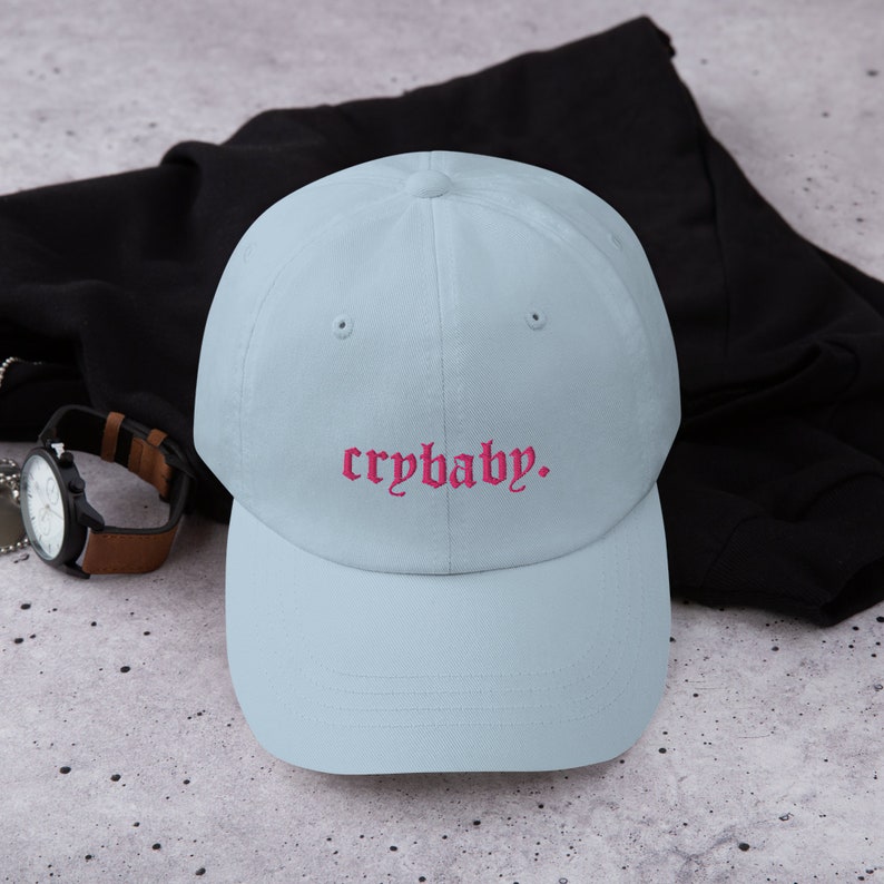 Puede incluir: Una gorra de b&eacute;isbol azul claro con el texto "crybaby" bordado en rosa.