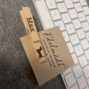 May include: A light-colored wooden clothespin featuring the name Max and a black silhouette of a dachshund, clipping a square tan card printed with the text Edelm&auml;dli personalisierte Lieblingsst&uuml;cke & zauberhafte Geschenke in black ink.