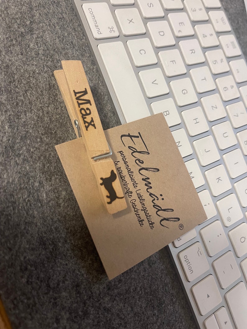 May include: A light-colored wooden clothespin featuring the name Max and a black silhouette of a dachshund, clipping a square tan card printed with the text Edelm&auml;dli personalisierte Lieblingsst&uuml;cke & zauberhafte Geschenke in black ink.
