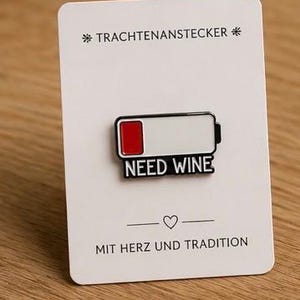Könnte beinhalten: Ein Metall-Emaille-Pin in Form eines Akku-Symbols mit rotem Segment und dem Text NEED WINE, befestigt auf einer weißen Trägerkarte mit dem Text TRACHTENANSTECKER oben und MIT HERZ UND TRADITION mit einem Herzsymbol am unteren Rand.