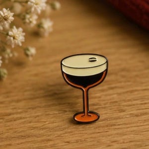 Könnte beinhalten: Eine Metall-Emaille-Anstecknadel in Form eines Cocktailglases, gefüllt mit einer dunkelbraunen Flüssigkeit, bedeckt mit einer dicken cremefarbenen Schaumschicht und einer mittig platzierten Kaffeebohne, mit schwarzer Umrandung und orangefarbenem Fuß.