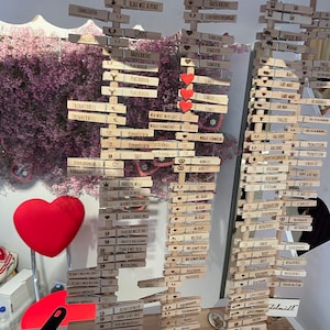 May include: Three vertical wooden displays holding numerous light-colored wooden clothespins, each engraved with various German phrases and small icons like hearts, crowns, and pretzels. Three red heart-shaped cutouts are attached to the center display.
