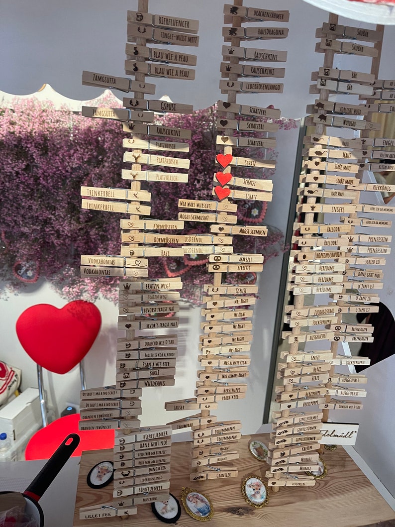 May include: Three vertical wooden displays holding numerous light-colored wooden clothespins, each engraved with various German phrases and small icons like hearts, crowns, and pretzels. Three red heart-shaped cutouts are attached to the center display.